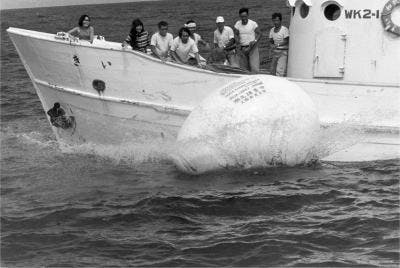 Voyage: Happening In An Egg by The Play, 1968. [A black and white photograph of the boat passing the floating egg structure. The people on the boat look at it from the deck.] 