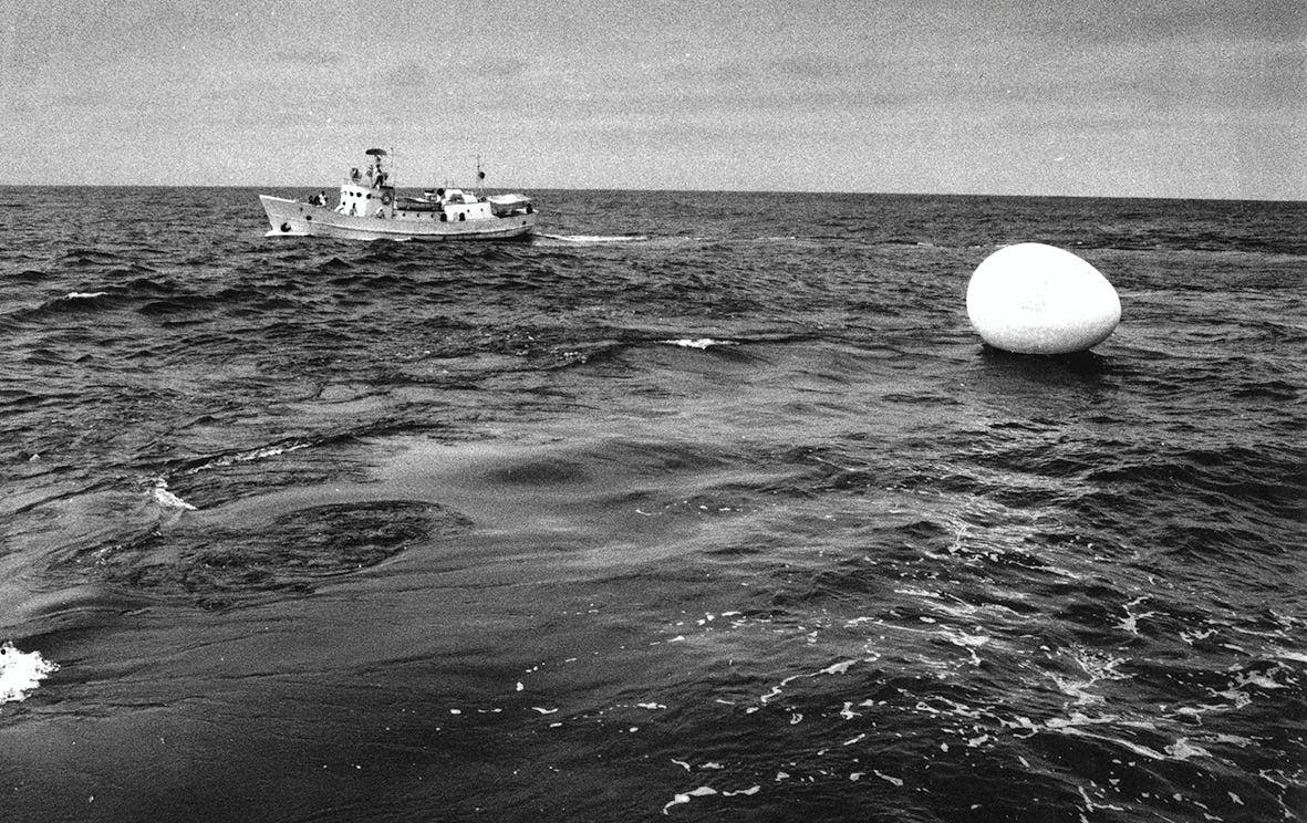 Voyage: Happening In An Egg by The Play, 1968. [A black and white photograph of the ocean, a boat in the background, and the egg sculpture floating on the water in the foreground.]