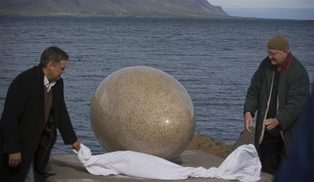 Eggin í Gleðivík (The Eggs of Merry Bay) by Sigurður Guðmundsson, 2009. [Two men pull a white sheet off a large granite egg, as if revealing it for the first time. The ocean can be seen in the background.]