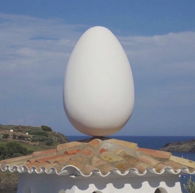 Salvador Dalí House in Portlligat, Spain. [A circular terracotta roof with an enormous white egg sitting atop it, as if balancing in place. Beyond the roof is the ocean and a bright blue sky.]