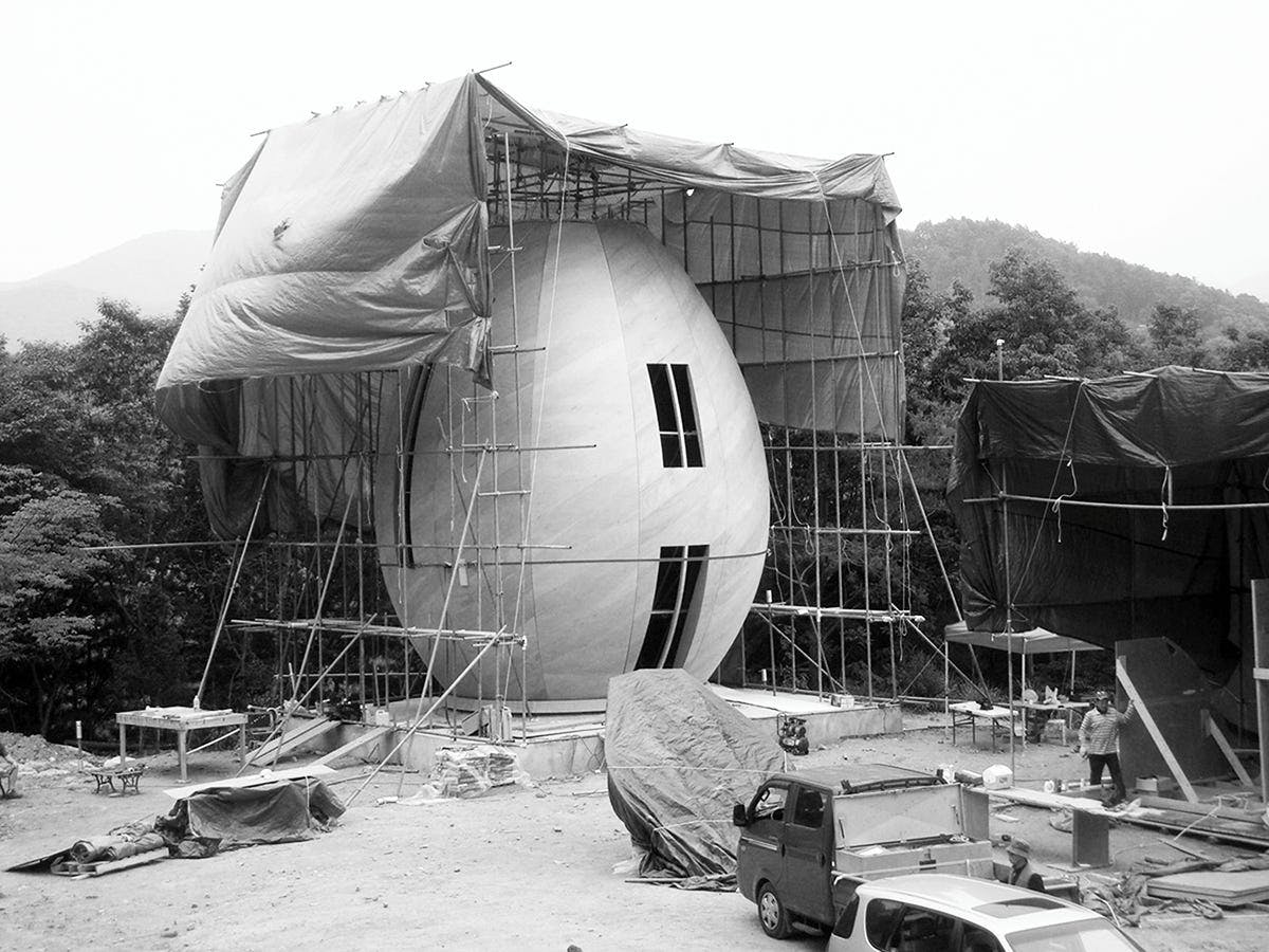 Egg Chapel by Andrew MacNair, 2012. [A black and white photograph showing the construction of a egg-shaped structure. Scaffolding is set up around it, and a window and door are cut out from the front.]