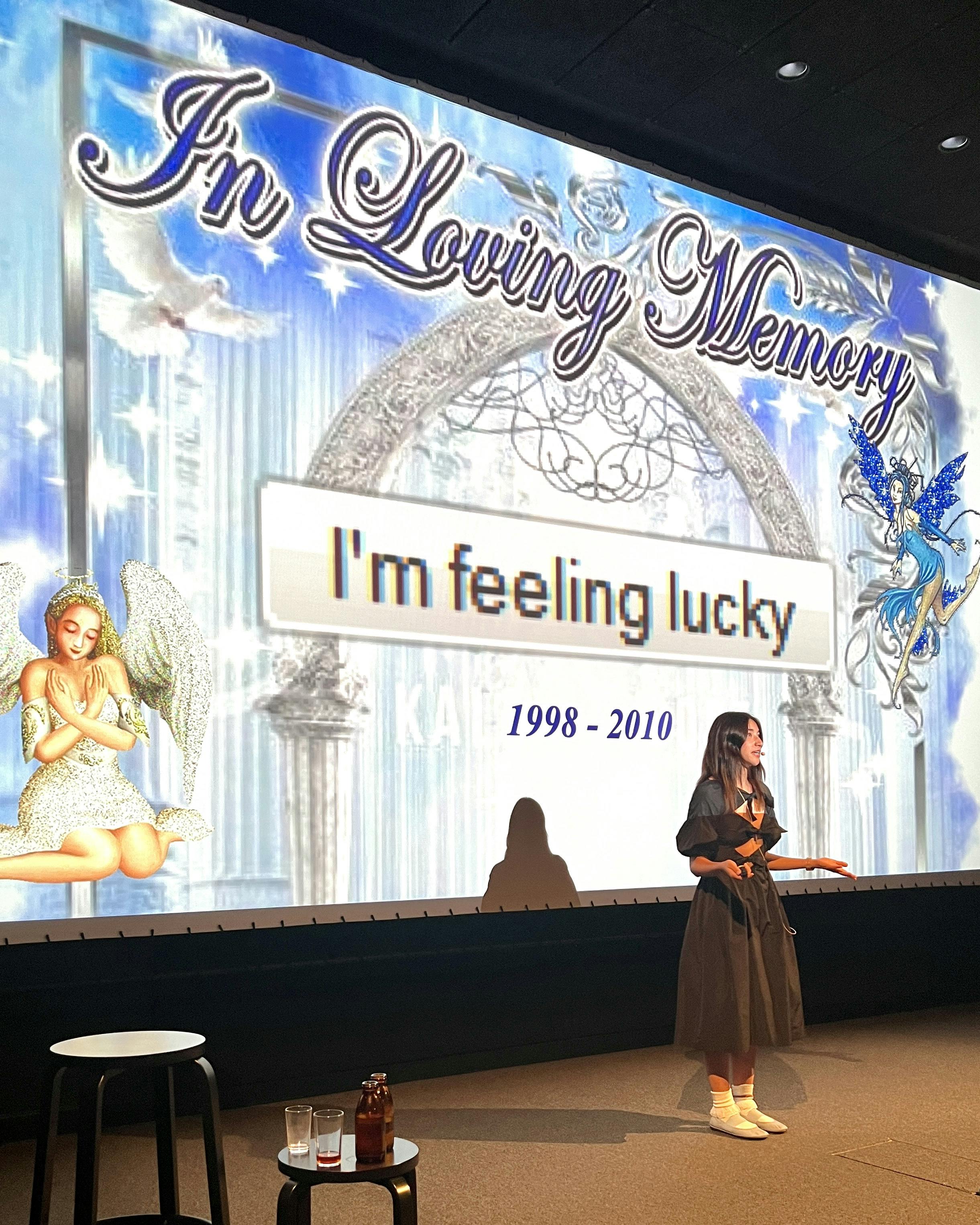 Maya giving the eulogy at Naive Yearly. [Maya stands before a giant screen that reads “In Loving Memory: I’m feeling lucky 1998-2010” with heavenly blue background, angels, doves, and a shimmering archway.]