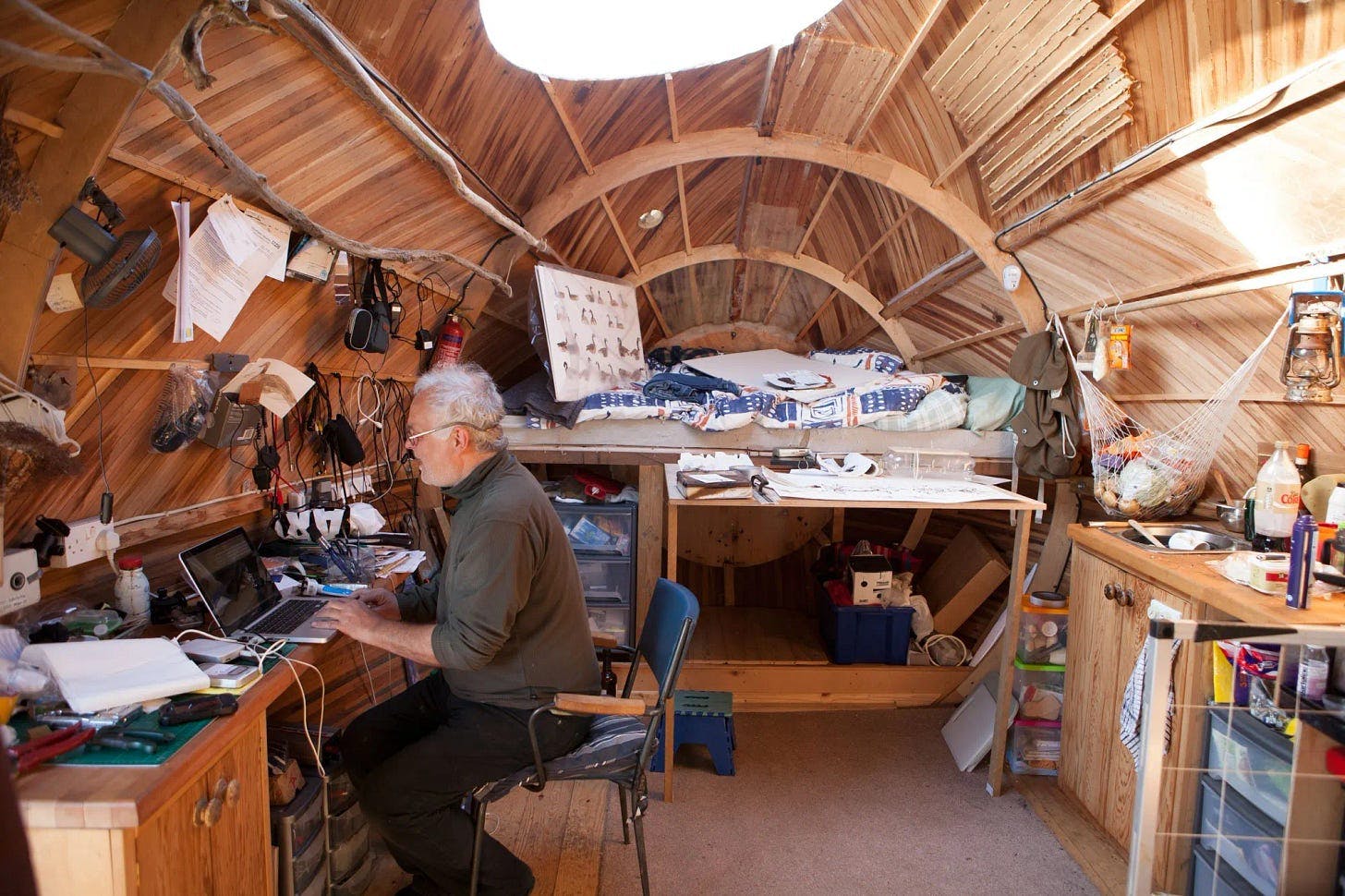 Exbury Egg, by Stephen Turner, 2013. [A man sits inside a homey oval wooden structure that almost looks like the hull of a ship. He works from a laptop on a desk strewn with stuff. Behind him is a kitchenette and beyond him looks like a living area.]